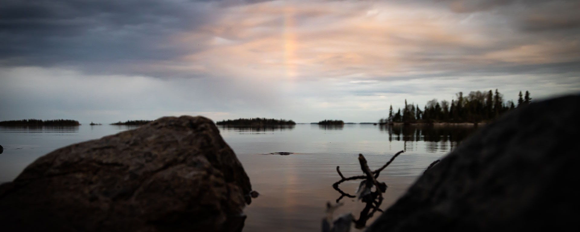 Dark clouds stretch out over a calm but Stoney lake a rainbow can be seen in the center.