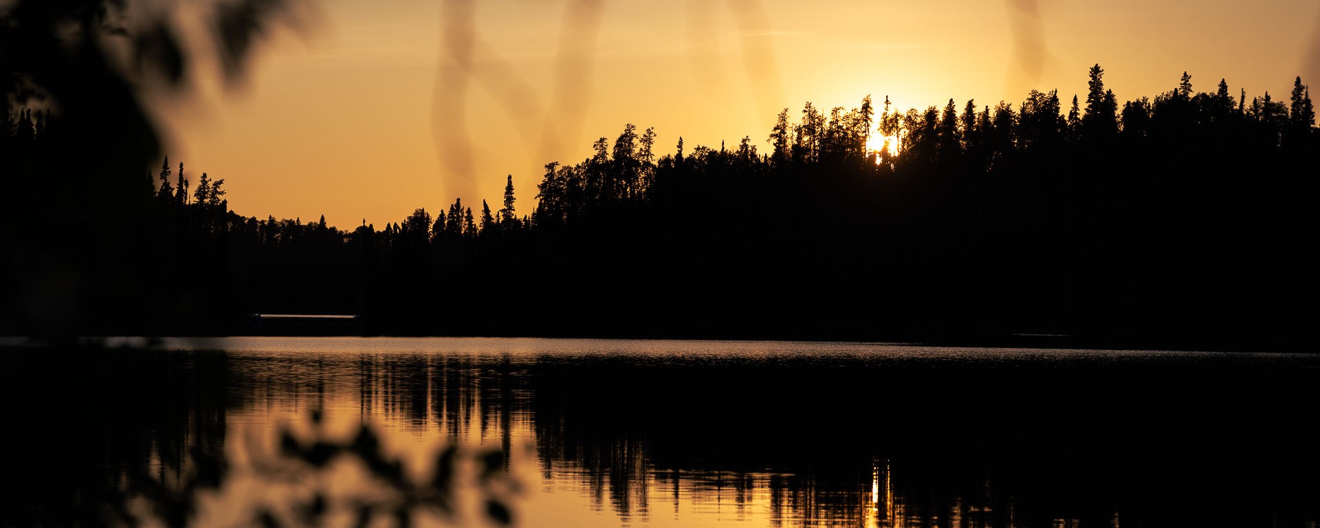 Sun falling behind trees lining a calm lake.